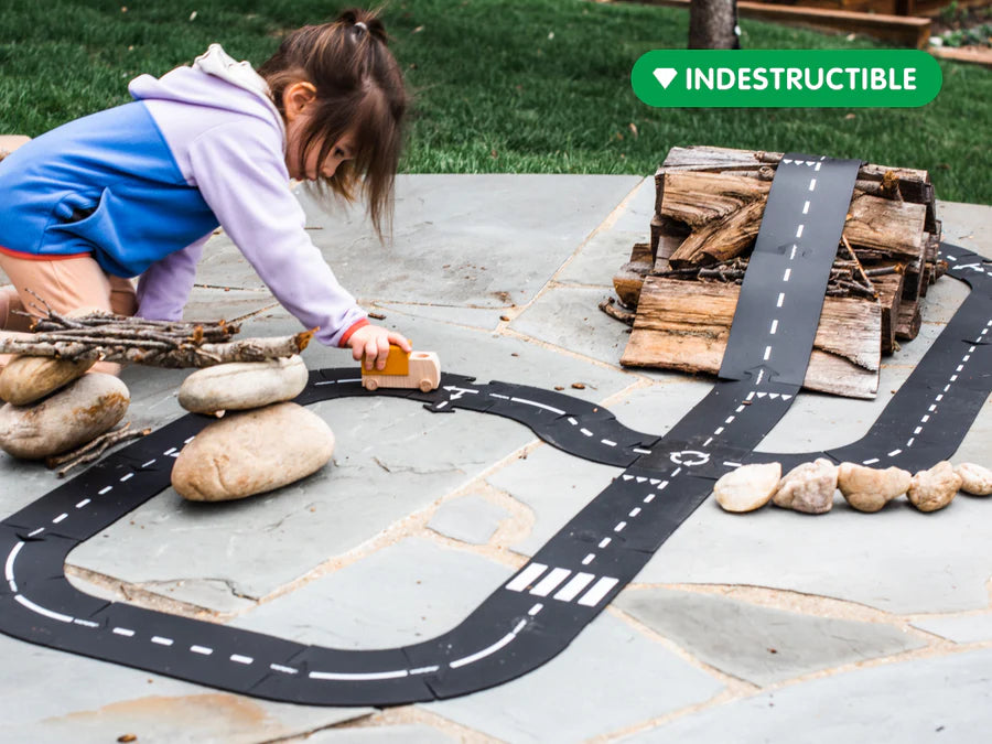 Child playing with a road made of black tape on a stone surface, with 'Indestructible' branding in the corner.