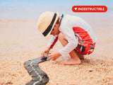 Child playing with toy cars on a sandy beach, wearing a hat and colorful outfit.